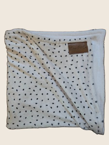 A folded plush fleece blanket in cream with black polka dots, featuring a brown faux-leather brand patch on the reverse side.
