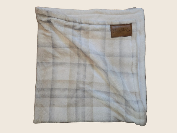 A folded plush throw blanket with a beige and white plaid pattern, featuring a small brown leather brand patch that reads 'Dunage' on a white background.