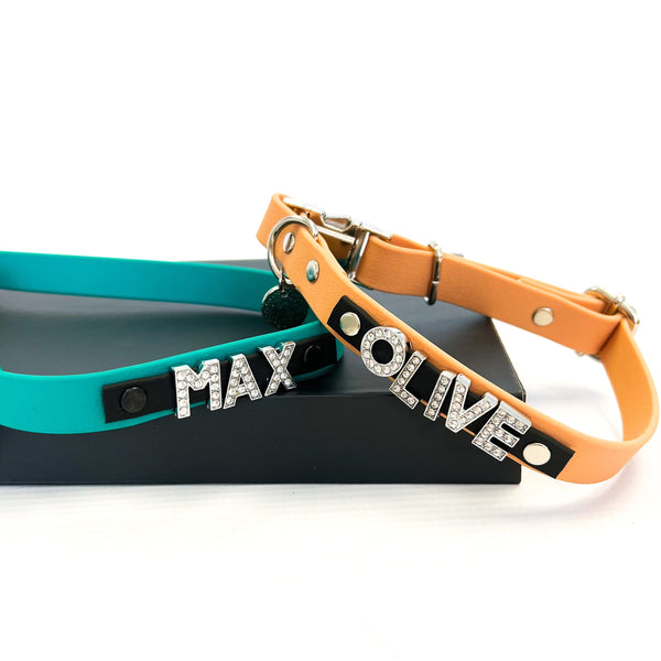 Two personalized dog collars with diamond charm letters spelling out 'MAX OLIVE' in diamonds, one in blue and the other in orange, displayed on a black box.